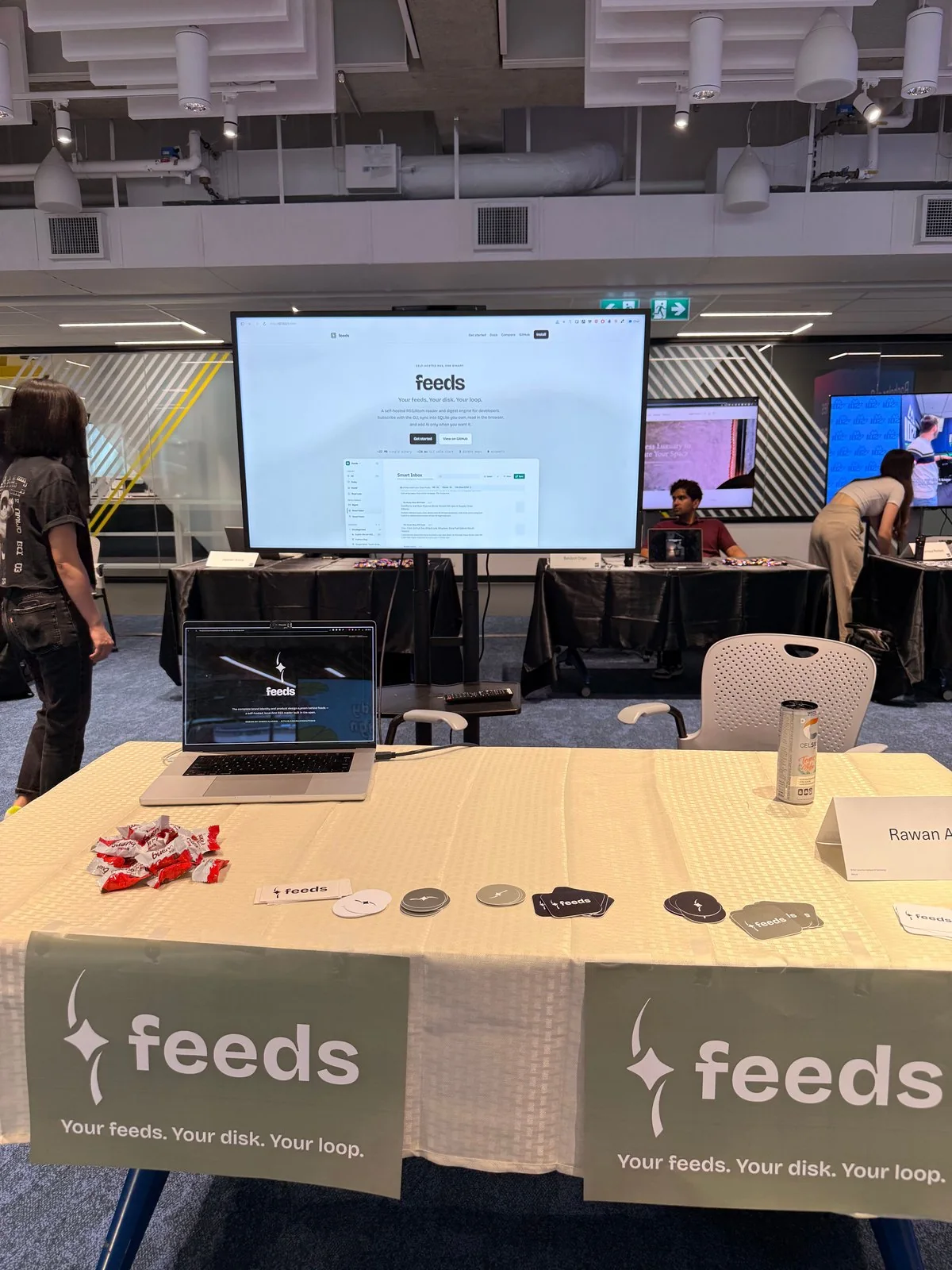 The Feeds booth at the showcase — two printed table runners carrying the mark and tagline, die-cut stickers on the table, a laptop looping the brand-identity reel, and the live product site on the screen behind.